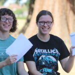 Two smiling friends with glasses holding white envelopes outdoors, sunlight and a tree behind them.