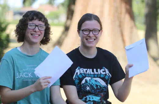 Two smiling friends with glasses holding white envelopes outdoors, sunlight and a tree behind them.