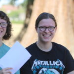 Two smiling friends with glasses holding white envelopes outdoors, sunlight and a tree behind them.
