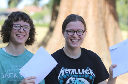 Two smiling friends with glasses holding white envelopes outdoors, sunlight and a tree behind them.